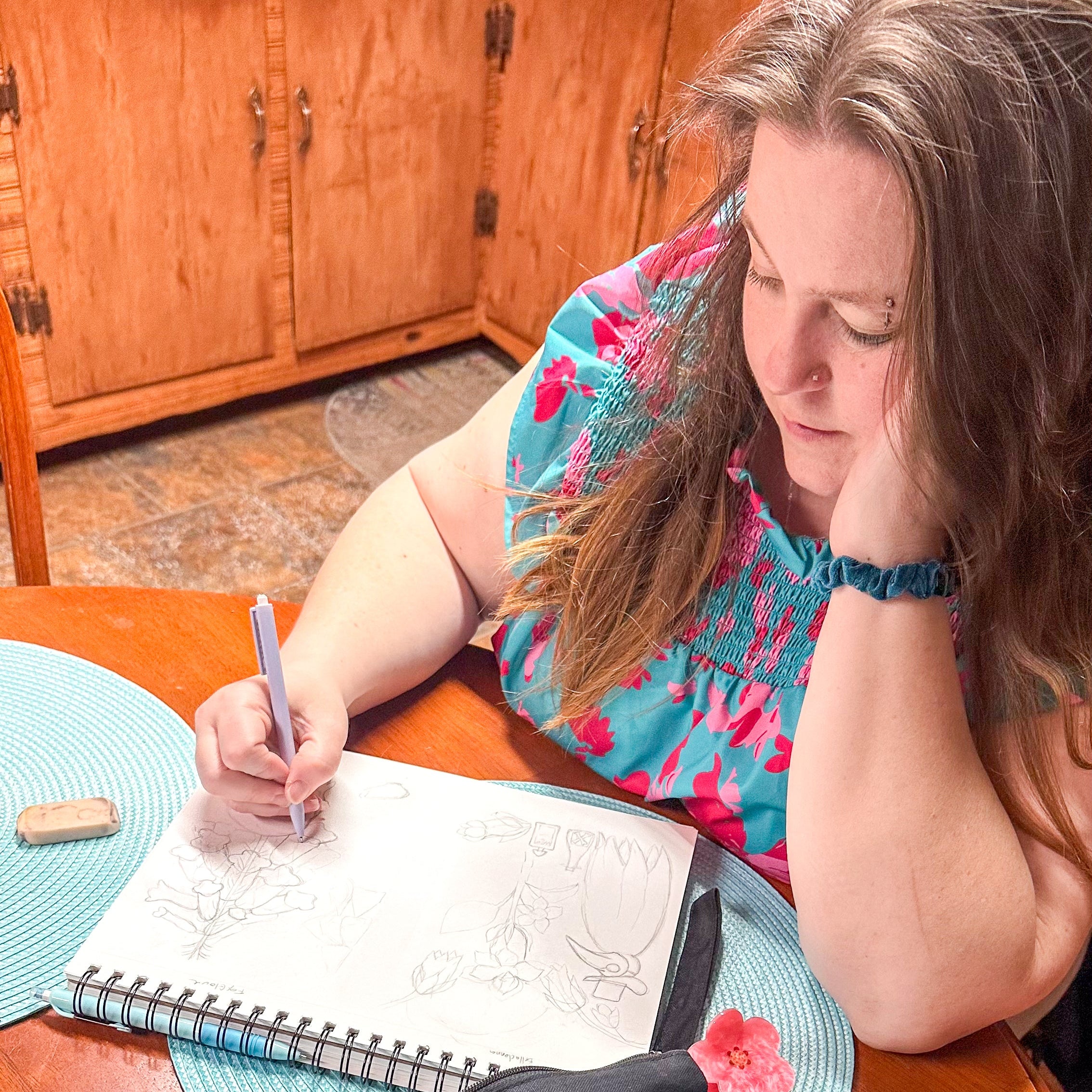 Woman sitting at a table with a notebook and pen, next to a black pouch with gold designs.