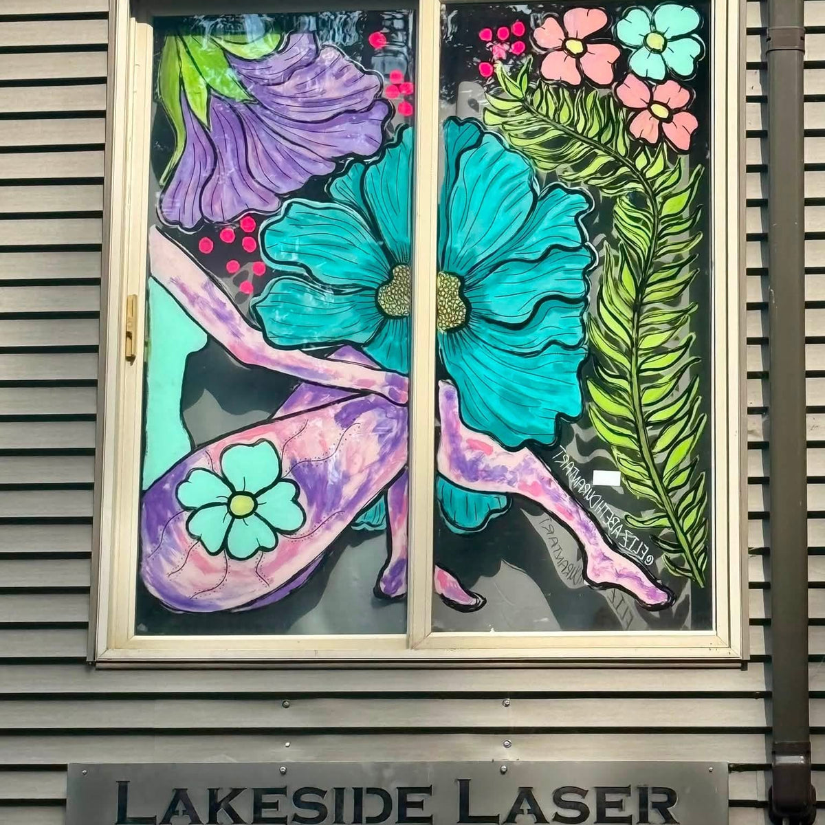 A decorative stained-glass style window on a gray shingled building, featuring large turquoise and purple flowers, pink blossoms, green leaves, and abstract curved lines. Below the window is a metal sign reading "LAKESIDE LASER" with "Est 2023" underneath.