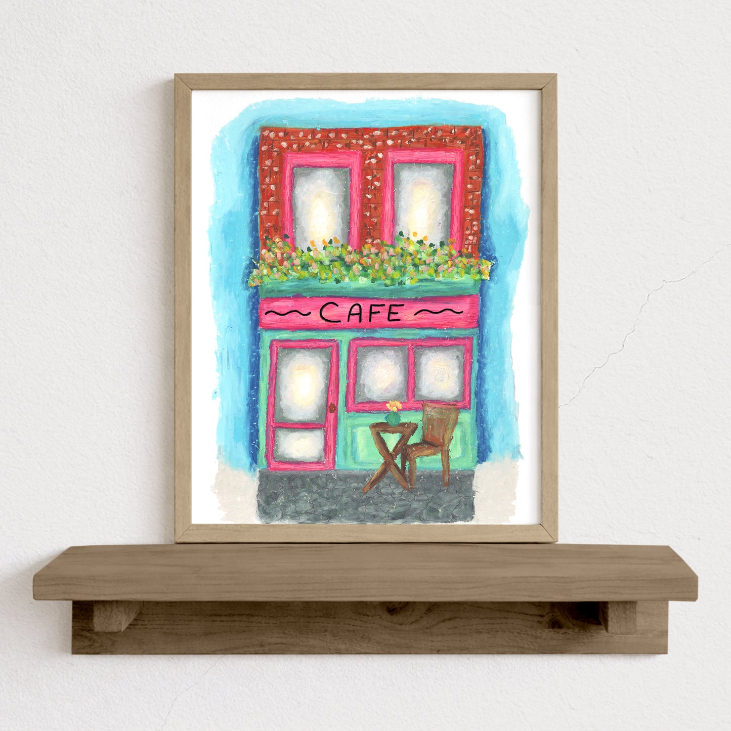 Framed artwork of a colorful cafe on a white wall with a wooden shelf.