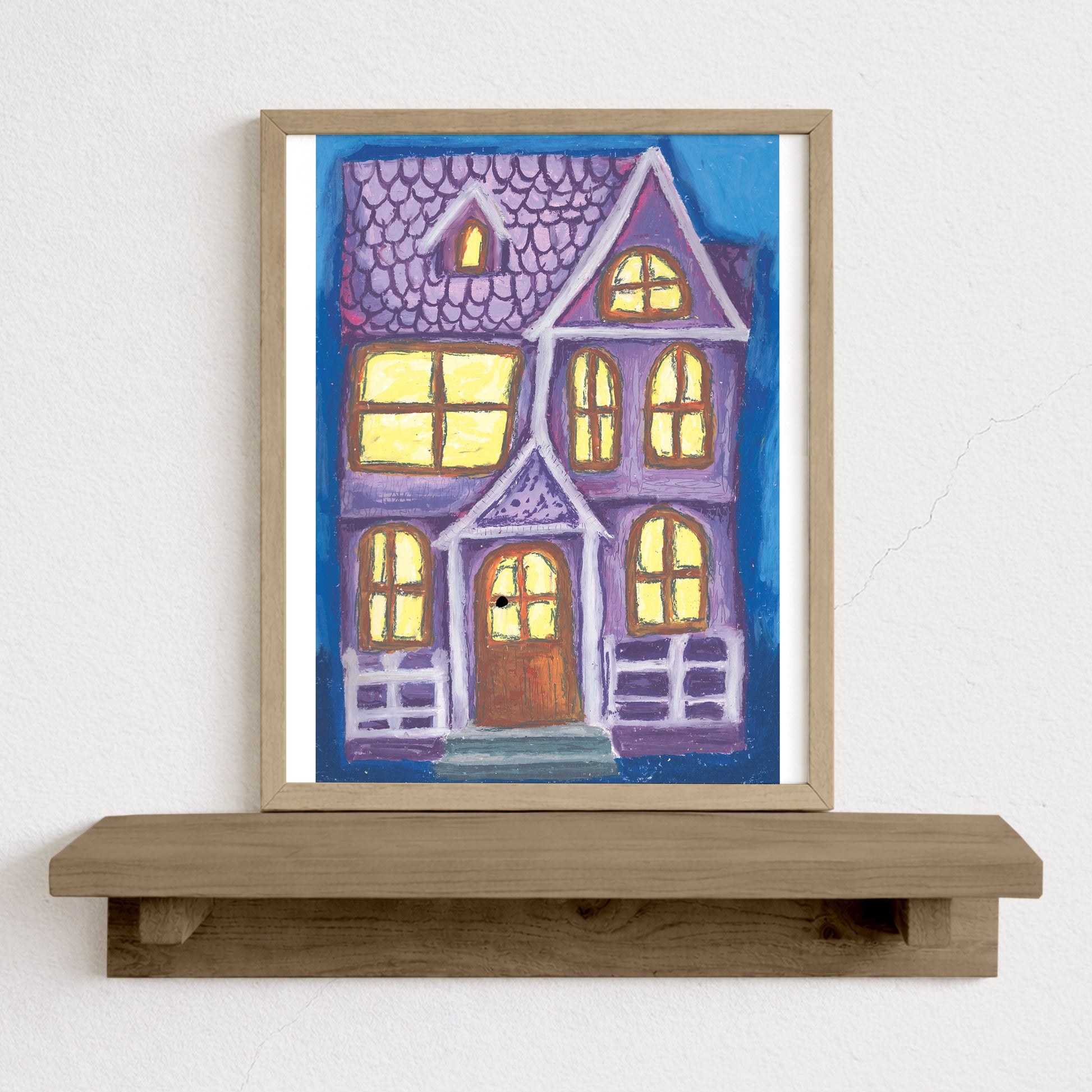 Framed artwork of a colorful house on a wooden shelf against a white wall