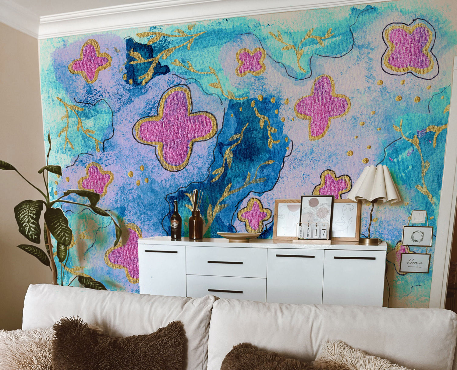 A styled living room featuring a large, vibrant abstract wall mural with blue, pink, and gold brushstrokes behind a white sofa with brown and beige textured pillows.